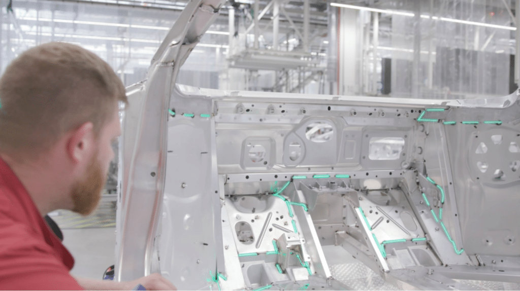 Ein Werker blickt in die Rohkarosserie, grüne Laserlinien projizieren Sollkonturen und Montagepositionen im Innenraum zur prozesssicheren Ausrichtung.