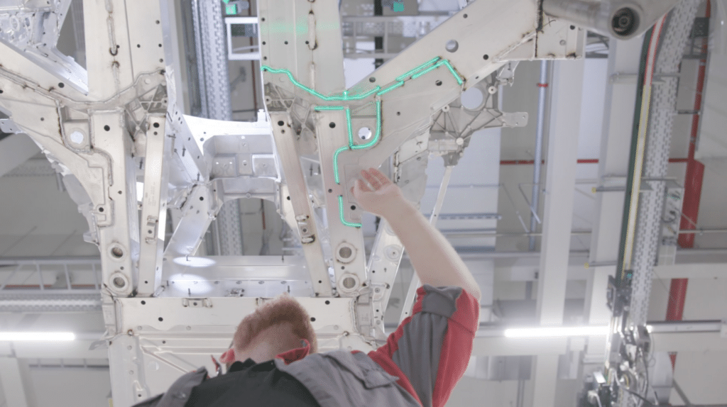 An operator beneath a body-in-white; green laser projection highlights contours/positions on a bodywork component for visual worker guidance.
