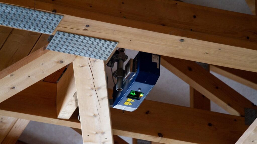 A laser projector supports the assembly of a nail plate truss, visible between timber beams and metal connectors.