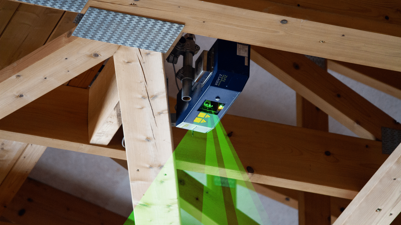 Der Z-LASER ZLP2 High Power Laserprojektor unter einem hölzernen Dachstuhl. Der Laserprojektor projiziert die CAD-Daten der Nagelbinder mit grünen Laserlinien.