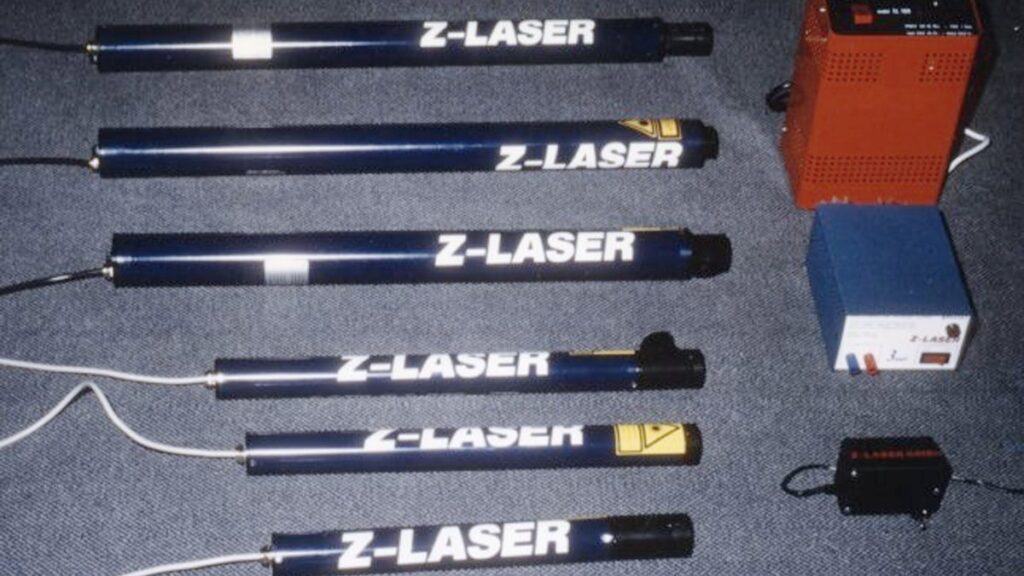 Mehrere frühe Z‑LASER Lasermodule mit „Z‑LASER“-Aufdruck liegen nebeneinander; daneben sind passende Netzteile/Steuergeräte zu sehen.