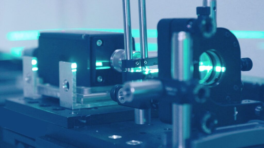 Laseraufbau im Labor: Optische Komponenten auf einer optischen Bank, sichtbar ist ein blaugrüner Laserstrahl zur Mess- und Entwicklungsarbeit.