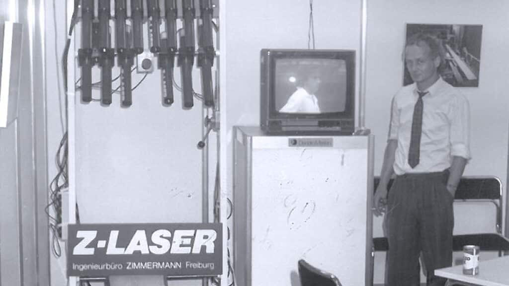 Schwarzweißaufnahme des Gründers von Z-LASER, Kurt-Michael Zimmermann. Zimmermann steht neben einem Fernseher und einem Setup von Lasern in seinem Ingenieursbüro in Freiburg.