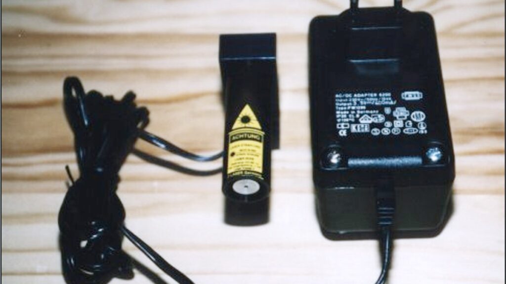 Modulares Lasermodul mit Laserwarnetikett neben einem Netzteil und Anschlusskabel auf Holzoberfläche.